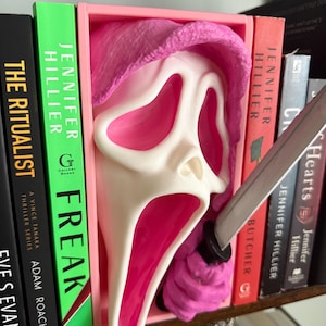 May include: A pink and white Ghostface mask bookend with a knife. The mask is set in a pink frame, with the knife appearing to pierce the mask. The bookend is placed between several books on a shelf.