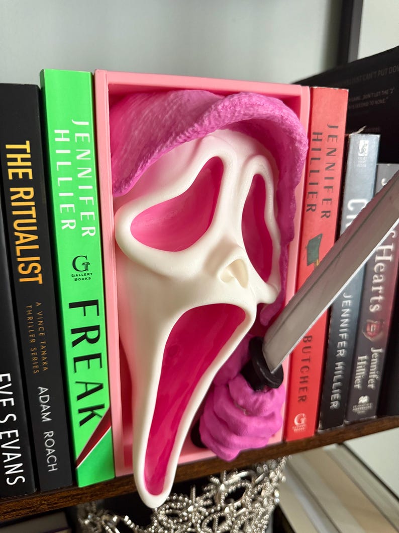 May include: A pink and white Ghostface mask bookend with a knife. The mask is set in a pink frame, with the knife appearing to pierce the mask. The bookend is placed between several books on a shelf.