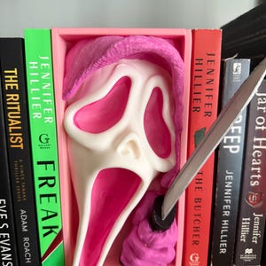 May include: A white and pink Scream mask with a pink hood and a silver knife, displayed between books on a shelf. The books have titles like "Freak" and "The Butcher" by Jennifer Hillier.