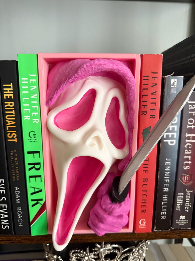 May include: A white and pink Scream mask with a pink hood and a silver knife, displayed between books on a shelf. The books have titles like "Freak" and "The Butcher" by Jennifer Hillier.