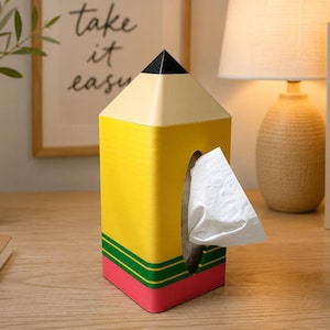 May include: A tissue box cover shaped like a yellow pencil with a black pointed tip, a cream-colored section, a yellow body, green stripes, and a pink base. A white tissue is pulled through the oval opening on the front of the pencil-shaped container.