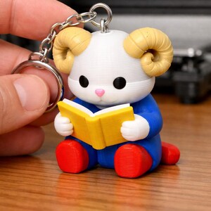 May include: A 3D-printed keychain featuring a cartoon ram reading a book. The ram has a white head, tan horns, black eyes, and a pink nose. It wears a blue outfit with red feet and holds a yellow book. The keychain is attached to a silver chain.
