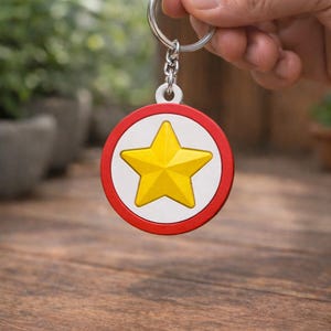 Pode incluir: Um porta-chaves redondo com um design de estrela amarela. A estrela está centrada num fundo branco, emoldurada por uma borda vermelha. O porta-chaves é preso a uma corrente e um anel prateados.