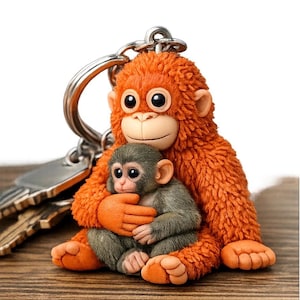 May include: A whimsical keychain featuring a mother monkey embracing a baby monkey. The mother monkey is orange with large eyes, and the baby monkey is gray. The keychain is attached to a silver key ring. The background is a wooden surface.