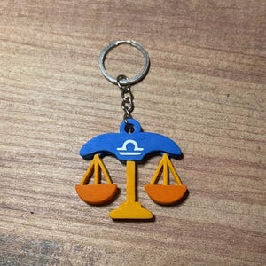Könnte beinhalten: Ein Schlüsselanhänger mit dem Sternzeichen Waage. Der Schlüsselanhänger hat ein blaues Waagen-Design mit orangefarbenen Gewichten und einem gelben Ständer. Der Schlüsselanhänger ist an einem silbernen Metallring befestigt.