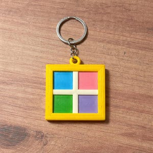Könnte beinhalten: Ein farbenfroher Schlüsselanhänger mit einem quadratischen Design, das in vier Quadrate in Blau, Rosa, Grün und Lila unterteilt ist, umrahmt von einem gelben Rahmen. Mit einem silbernen Metallring zum Befestigen an Schlüsseln.