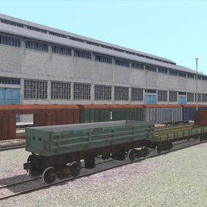 May include: A collection of freight train cars in various colors, including red, green, and blue, are parked on parallel tracks. The cars are in front of a large industrial building with multiple windows.