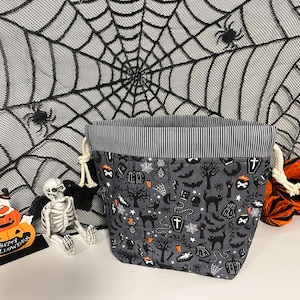 May include: A fabric storage basket featuring a grey background with a repeating Halloween pattern of black cats, bats, pumpkins, coffins, and spiders. The basket has a black and white pinstriped top border and thick cream-coloured drawstring cords.