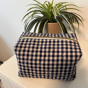 May include: A rectangular quilted pouch featuring a navy blue and white gingham check pattern. The bag has a structured shape with a top zip closure that includes a metallic gold-toned zip pull and white fabric trim along the zip line.