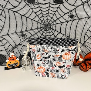 May include: A fabric drawstring bag featuring a white background with a pattern of orange pumpkins, black spiders, moths, and gothic structures. The top edge is trimmed with black fabric featuring small white polka dots and cream-coloured cords.