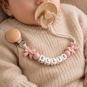 May include: A beige pacifier attached to a custom pacifier clip featuring a round wooden fastener, pink silicone beads, two pink bow-shaped accents, and white cube beads with black letters spelling out the name ELLIE on a light beige sweater.