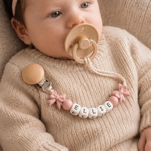 May include: A beige pacifier attached to a custom pacifier clip featuring a round wooden clip, pink silicone bow beads, and white cube beads spelling the name ELLIE in black letters, connected by a white cord to the beige silicone pacifier.