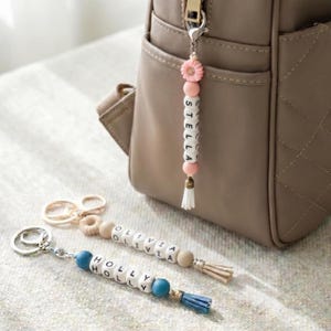 May include: Three personalized keychains featuring round beads and letter beads spelling the names Stella, Olivia, and Holly. Each keychain includes a metal ring, a small flower or textured bead, and a decorative tassel at the bottom end.