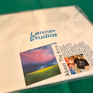 May include: A white card with the text "Leitner Studios" in blue lettering. The card also features a photo of a landscape with a pink and purple sky and a green field. The card also includes a smaller card with a photo of a man and the text "About The Artist" and a description of the artist.