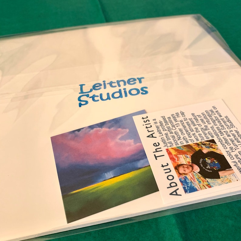 May include: A white card with the text "Leitner Studios" in blue lettering. The card also features a photo of a landscape with a pink and purple sky and a green field. The card also includes a smaller card with a photo of a man and the text "About The Artist" and a description of the artist.