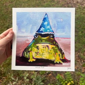 May include: A whimsical painting of a green frog wearing a blue wizard hat with white stars. The frog has yellow and green details, and the artwork is signed "jroc" with the title "Wizard Toad" below the frog.