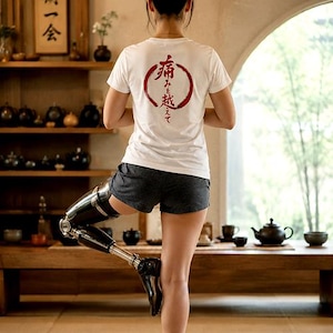 Könnte beinhalten: Eine Person in einer Yoga-Pose, trägt ein weißes T-Shirt mit japanischem Text in einem roten Kreis. Die Person hat ein Beinprothese. Der Hintergrund umfasst Holzregale mit Teesets und ein Fenster.