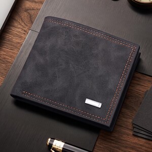 May include: A dark gray bi-fold wallet with a textured, weathered finish and contrasting brown stitching along the edges. The wallet features a small, rectangular silver-toned metal accent plate positioned on the lower right corner of the front cover.