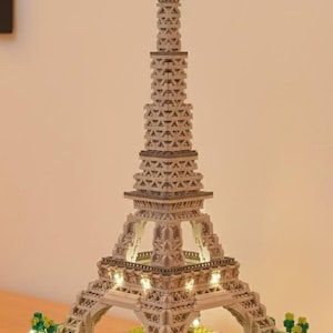 May include: A detailed model of the Eiffel Tower, constructed from interlocking blocks. The gray structure features intricate detailing, with lights illuminating the base and tower. A small red flag tops the tower. The model sits on a base with miniature trees.