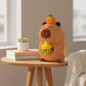 May include: A brown voxel-style capybara figurine with a pumpkin on its head and a yellow bubble tea cup. The figurine is on a wooden table next to a small succulent plant and books.