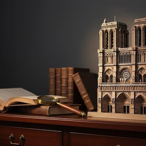 May include: A detailed wooden model of the Notre Dame Cathedral stands on a dark wood surface, next to an open book and a magnifying glass. Several antique books are stacked nearby. The cathedral model features intricate architectural details.