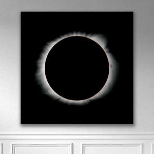Puede incluir: Impresión en lienzo cuadrada que representa un eclipse solar. La imagen presenta un gran círculo negro, la luna, rodeado por una brillante corona blanca sobre un fondo negro. La obra de arte está montada en una pared blanca.