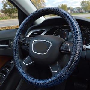 May include: A dark blue steering wheel cover featuring a glossy, embossed crocodile skin texture. The cover is fitted onto a black steering wheel with silver accents, positioned inside a vehicle interior with a view of a road through the windshield.