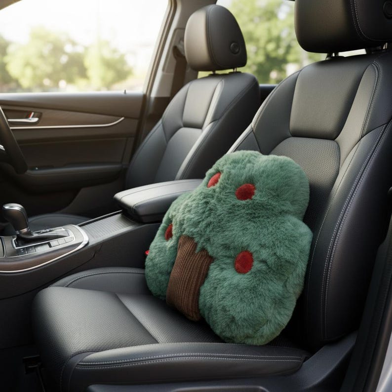 May include: A green plush tree-shaped pillow with red accents and a brown trunk sits on a black car seat. The pillow is designed to resemble a tree with a fluffy texture and is placed in the driver's seat.