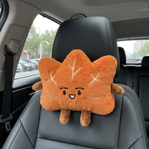 May include: A plush, orange, leaf-shaped car headrest pillow featuring an embroidered face with black eyes and a smiling mouth. The soft, fuzzy material includes stitched vein details on the surface, along with two small arms and two legs.