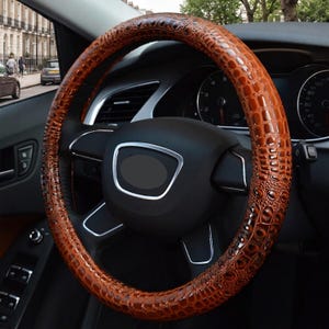 May include: A brown steering wheel cover with a textured crocodile skin pattern. The cover is fitted onto a black steering wheel with silver accents, featuring a circular shape and a glossy finish that highlights the embossed reptile scale design.