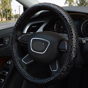 May include: A black steering wheel cover featuring a glossy, textured crocodile skin pattern. The cover is fitted onto a standard vehicle steering wheel with a central black hub, silver trim accents, and integrated control buttons on the spokes.