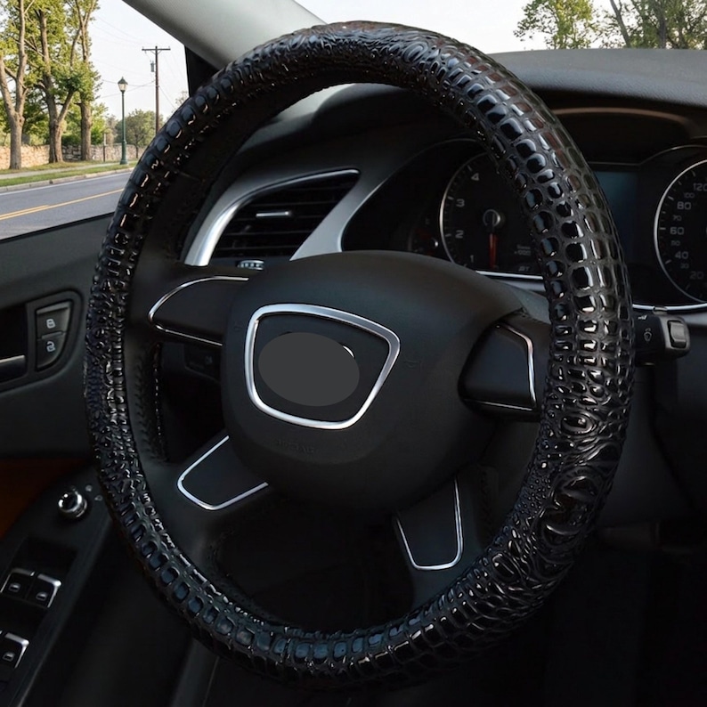 May include: A black steering wheel cover featuring a glossy, textured crocodile skin pattern. The cover is fitted onto a standard vehicle steering wheel with a central black hub, silver trim accents, and integrated control buttons on the spokes.