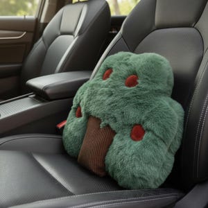 May include: A green plush tree-shaped pillow with red accents and a brown trunk sits on a black leather car seat. The pillow has a soft, furry texture and is designed for comfort and support. The car interior is visible in the background.