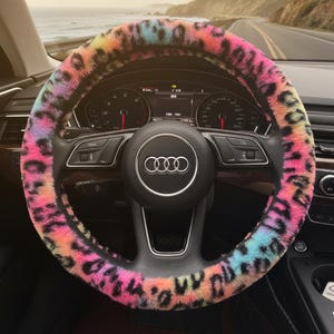 May include: A colorful, faux fur steering wheel cover with a leopard print design. The cover features a vibrant mix of pink, yellow, blue, and orange hues. The cover is installed on a black steering wheel in a car.