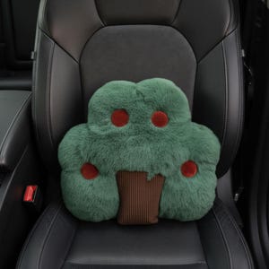 May include: A green, tree-shaped car seat pillow with red accents and a brown trunk. The pillow is made of a soft, plush material and is designed to provide comfort and support while driving. The pillow is placed on a black car seat.