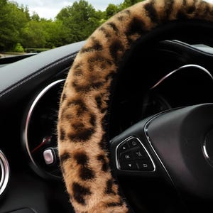 Pode incluir: Um volante de carro com uma capa com estampa de leopardo marrom e preto. A capa &eacute; feita de um material macio e felpudo. O volante &eacute; preto com detalhes prateados e bot&otilde;es de controle.
