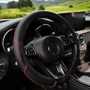 Puede incluir: Funda de volante negra y roja con un patrón de diamantes. La funda está cosida con hilo rojo y tiene un diseño deportivo. El volante está en el interior de un coche con vistas a un paisaje verde.