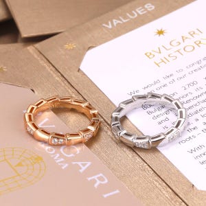 Puede incluir: Dos anillos con un diseño segmentado similar a una serpiente. Un anillo es de oro rosa y el otro de metal plateado. Ambos anillos presentan pequeñas piedras claras y brillantes engastadas en segmentos alternos a lo largo de la banda.