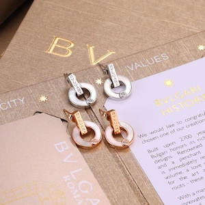 May include: Four drop earrings featuring a circular pendant design with the BVLGARI logo engraved on the vertical bar. Two earrings are silver-toned with white circular insets, and two are rose gold-toned with white circular insets, each with a small stone.