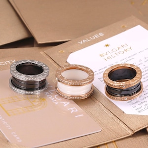 Könnte beinhalten: Drei Bvlgari-Ringe mit zylindrischem Design und eingraviertem Markennamen. Ein Ring ist silberfarben mit schwarzer Mitte und Diamanten, einer ist roségold mit weißer Mitte, einer ist roségold mit schwarzer Mitte und Diamanten.