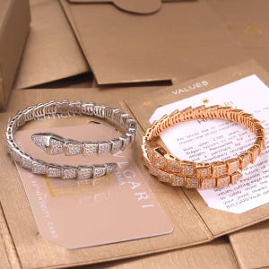 Puede incluir: Dos pulseras en forma de serpiente enrollada, una en metal plateado y otra en metal tono oro rosa, ambas con piedras claras en pavé. Cada pulsera descansa sobre una tarjeta de marca con texto que incluye BVLGARI y VALUES.