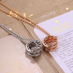 May include: Two necklaces featuring coiled, snake-shaped pendants with pavé-set diamonds. One pendant is crafted from rose gold, and the other is made of silver-toned metal. Each pendant hangs from a matching delicate chain resting on a document.