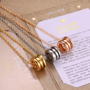 May include: Three necklaces featuring cylindrical pendants with ribbed detailing. One pendant is gold-toned, one is silver-toned, and one is rose gold-toned. Each pendant is suspended from a matching delicate metal chain link necklace.