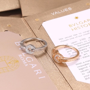 Puede incluir: Dos anillos, uno de metal plateado y otro de metal en tono oro rosa, cada uno con una pieza central circular incrustada con pequeñas piedras claras y el texto BVLGARI grabado en la banda, sobre documentación de papel de la marca.