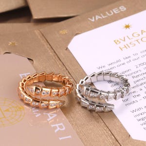 Pode incluir: Dois anéis em forma de serpente enrolada, um em ouro rosa e outro em metal prateado, ambos com corpos segmentados incrustados com pequenas pedras claras. Os anéis estão sobre cartões de papel com o texto visível BVLGARI.