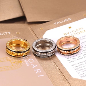 May include: Three Bvlgari rings arranged in a row, featuring one yellow gold, one silver-toned, and one rose gold band. Each ring has a central textured band with a repeating geometric pattern and the brand name engraved on the top and bottom edges.