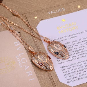 Puede incluir: Dos collares de oro rosa con colgantes en forma de cabeza de serpiente incrustados con diamantes pavé transparentes. Un colgante tiene ojos de piedra azul oscuro y el otro ojos de piedra roja. Las cadenas tienen eslabones hexagonales sobre papel.
