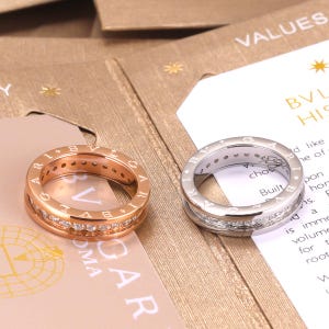 Puede incluir: Dos anillos, uno en oro rosa y otro en plata, cada uno con una banda central de pequeñas piedras claras y el texto BVLGARI grabado en los bordes exteriores. Los anillos están colocados sobre tarjetas de papel impresas con texto visible.