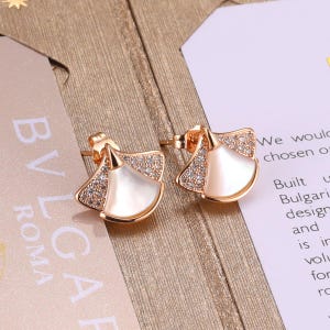 May include: A pair of rose gold-toned stud earrings shaped like fans, featuring a central white inlay and small clear stones along the edges. The earrings have a post-back closure and are displayed on a light-colored surface with visible text.