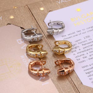 Könnte beinhalten: Drei Paar Creolen in Silber-, Gold- und Roségoldtönen, jeweils mit einem segmentierten, schlangenartigen Design, das mit kleinen, klaren, funkelnden Steinen besetzt ist. Die Ohrringe sind in einer vertikalen Reihe auf einer Papierkarte angeordnet.